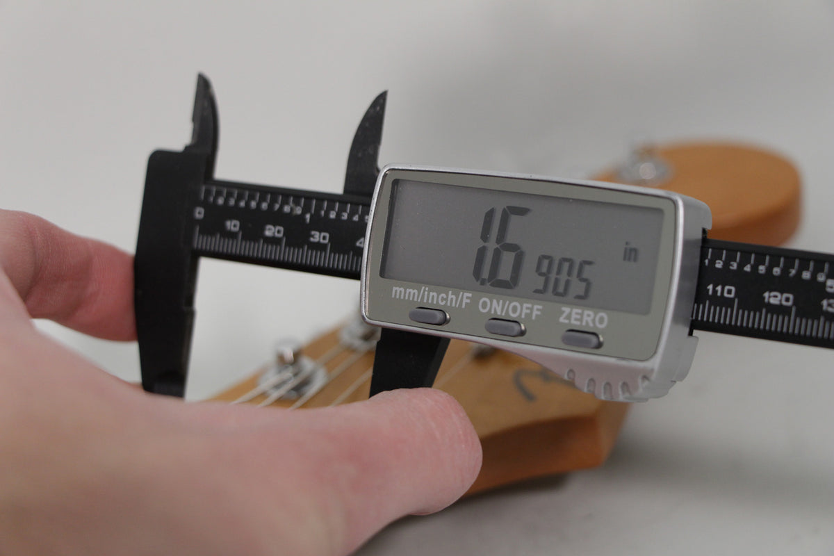 A hand holds a digital caliper measuring the guitar's nut, displaying "1.6905 in" on its screen, with the headstock visible.