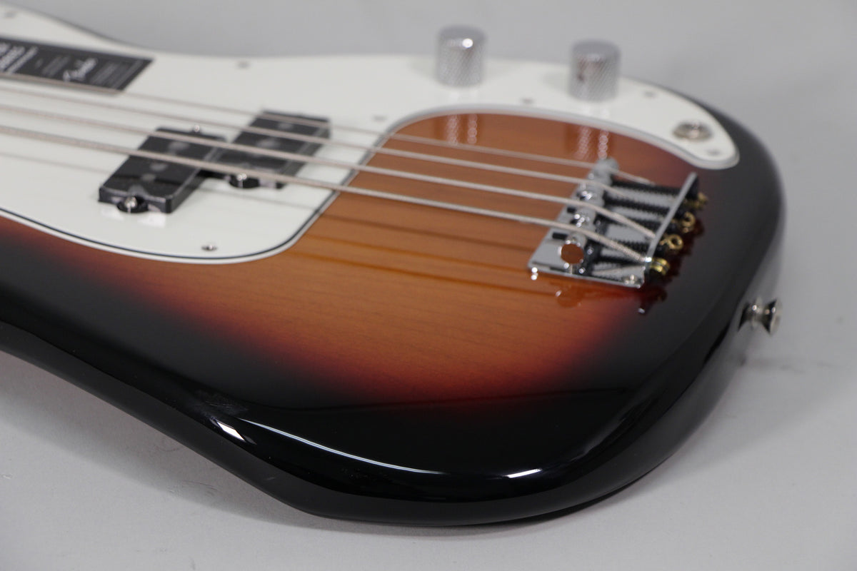 Close-up of a sunburst bass guitar body, showing the glossy finish, white pickguard, split pickup, chrome bridge, and two control knobs.