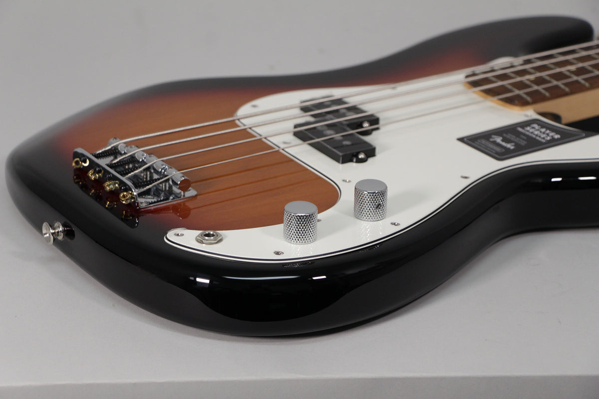 The bass body, in a glossy sunburst finish with a white pickguard, is angled from the lower bout, showing the bridge and two chrome knurled control knobs.