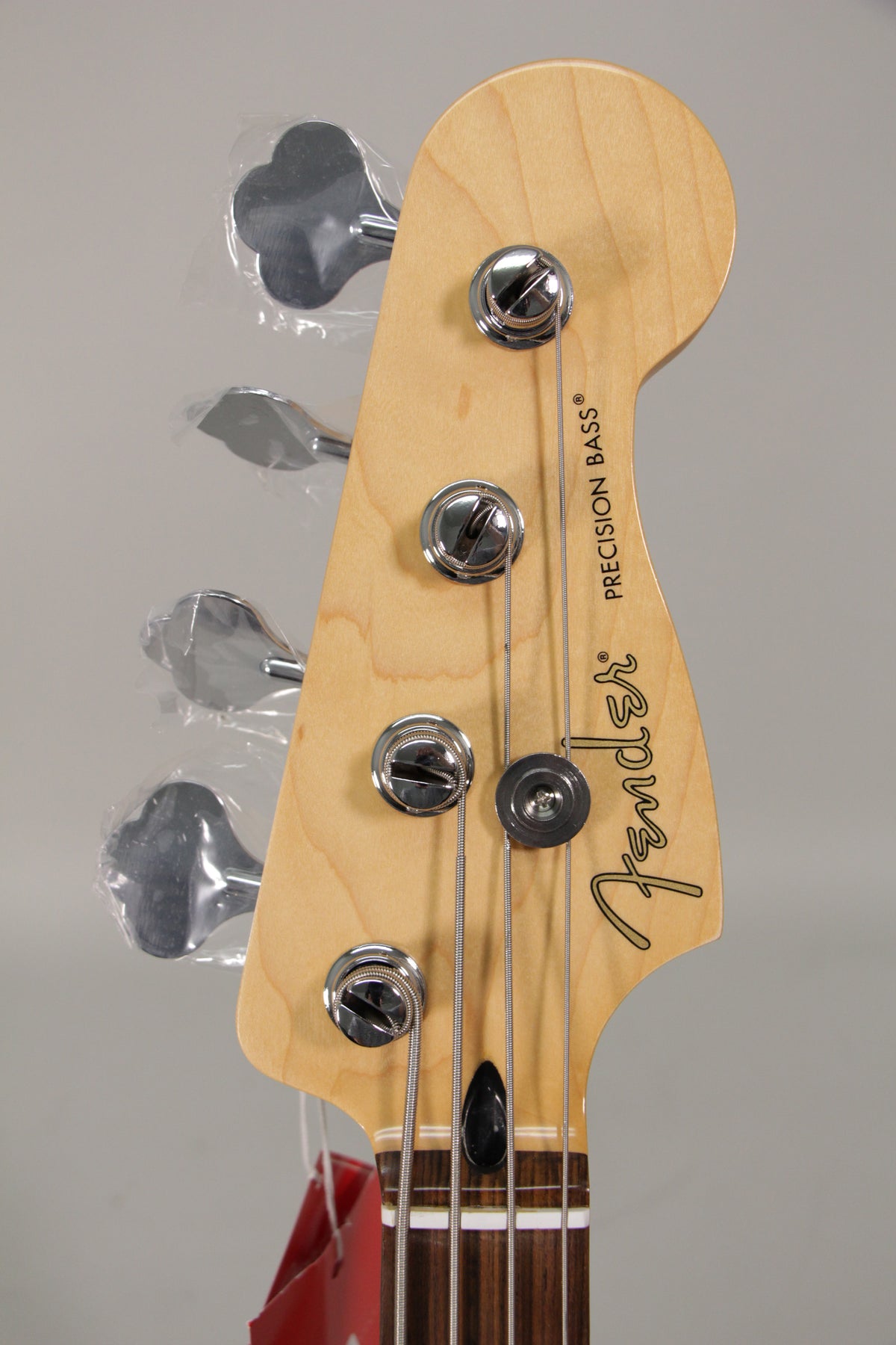 Close-up of the bass headstock, showing the light wood finish, four chrome tuning pegs with plastic still on, and the top of the dark fretboard.