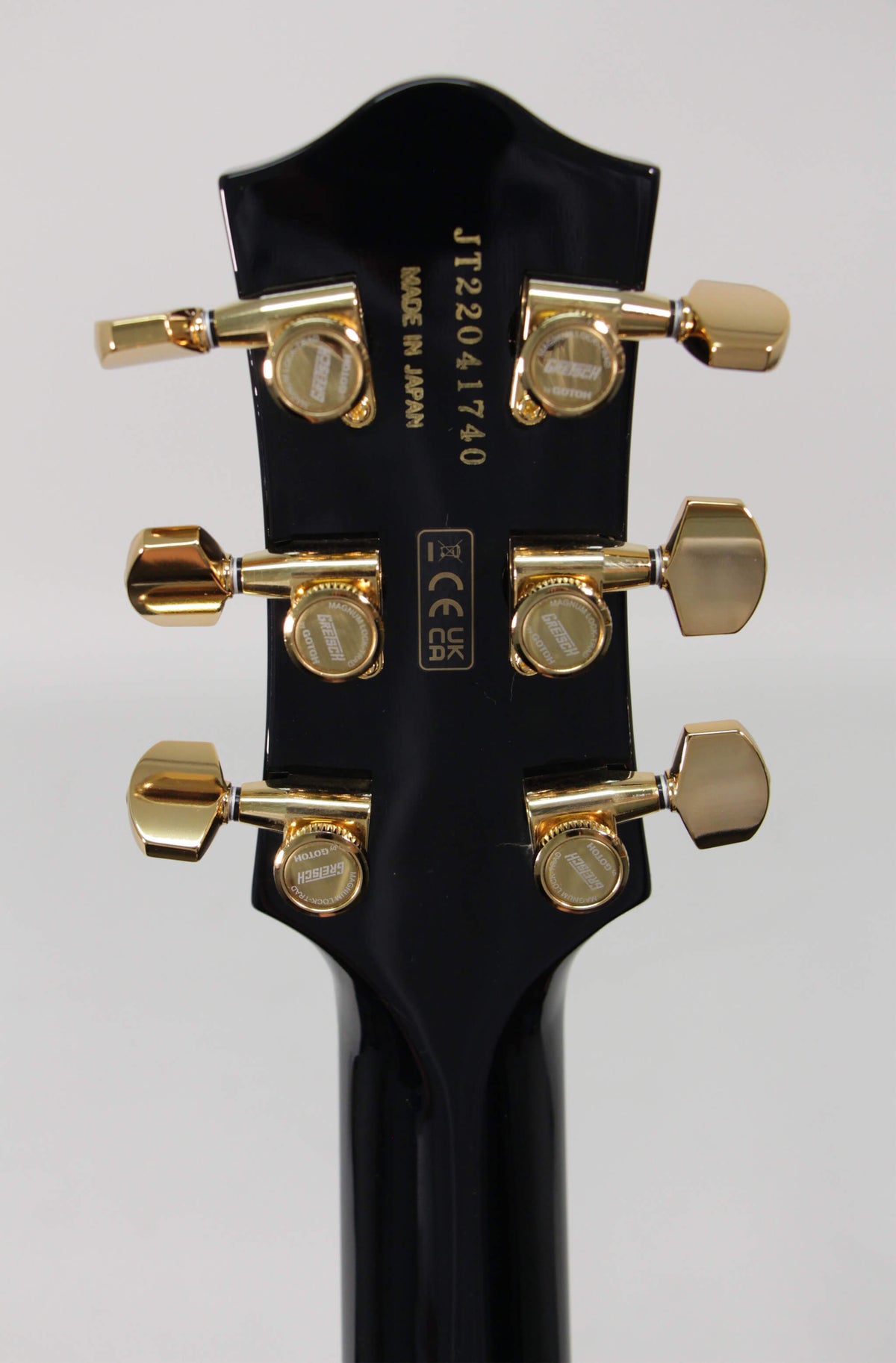 The guitar's glossy black headstock, viewed from the rear, features six gold tuning machines with rectangular buttons and visible serial number.