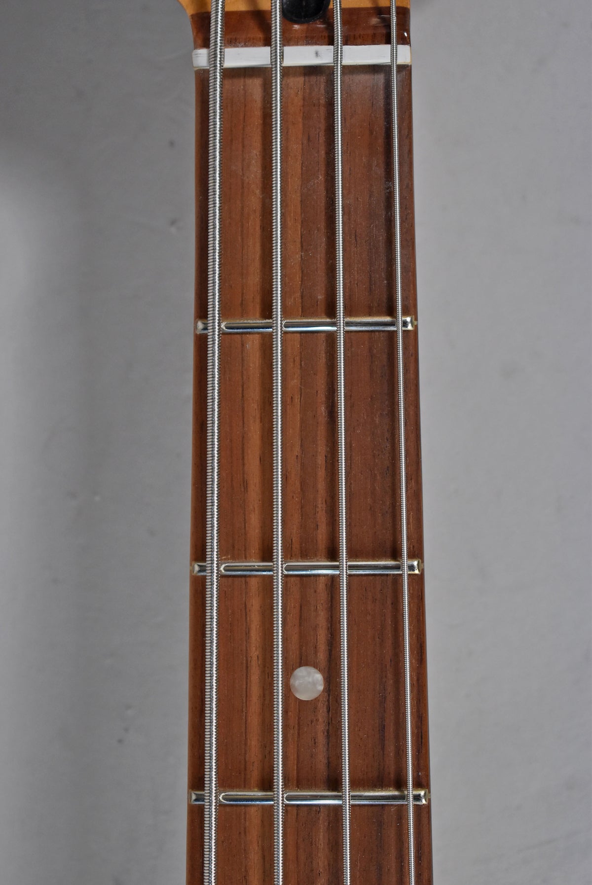 Close-up of the bass guitar's dark brown fretboard with four strings, silver frets, and a single round white fret marker.