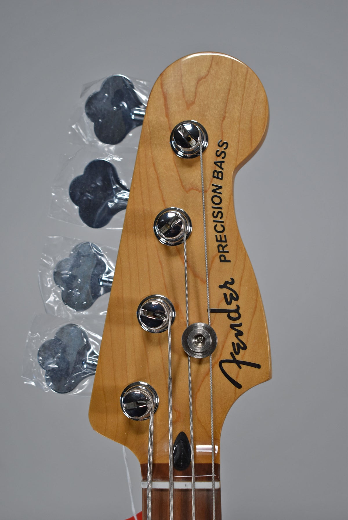 The bass headstock, angled slightly left, shows a light wood finish, chrome tuners with plastic still on, and a black string tree.