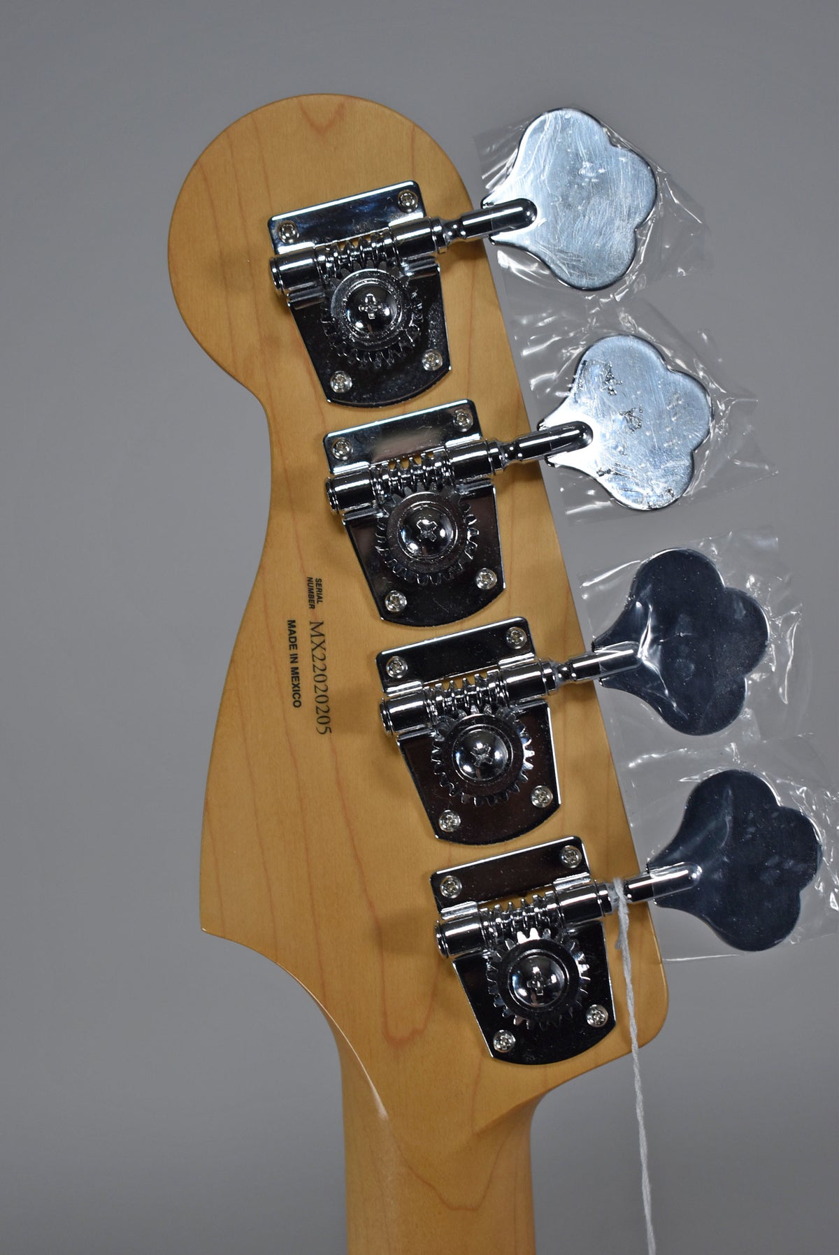 The bass headstock, light wood with a satin finish, is viewed from behind, showing four chrome tuners with plastic-wrapped keys.