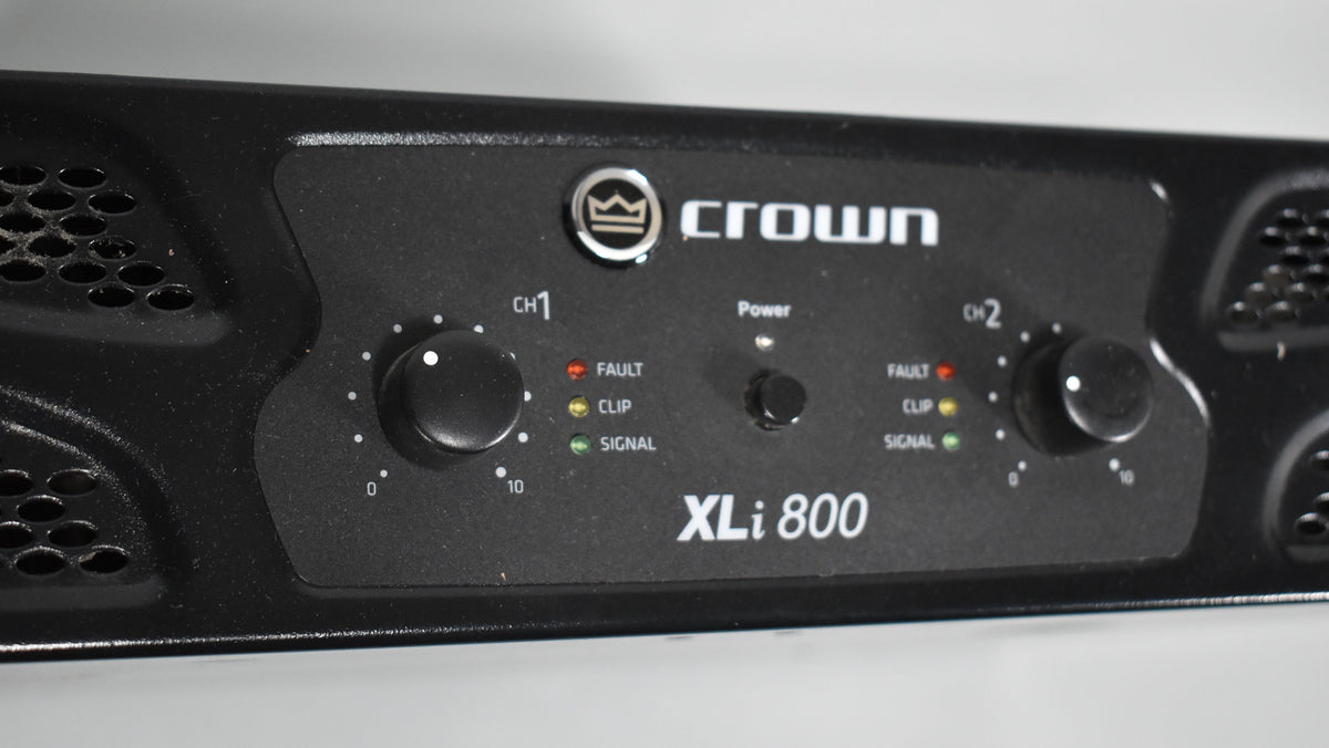 Close-up of the black amplifier's front panel, showing two large channel knobs, a power button, and LED indicators for fault, clip, and signal.
