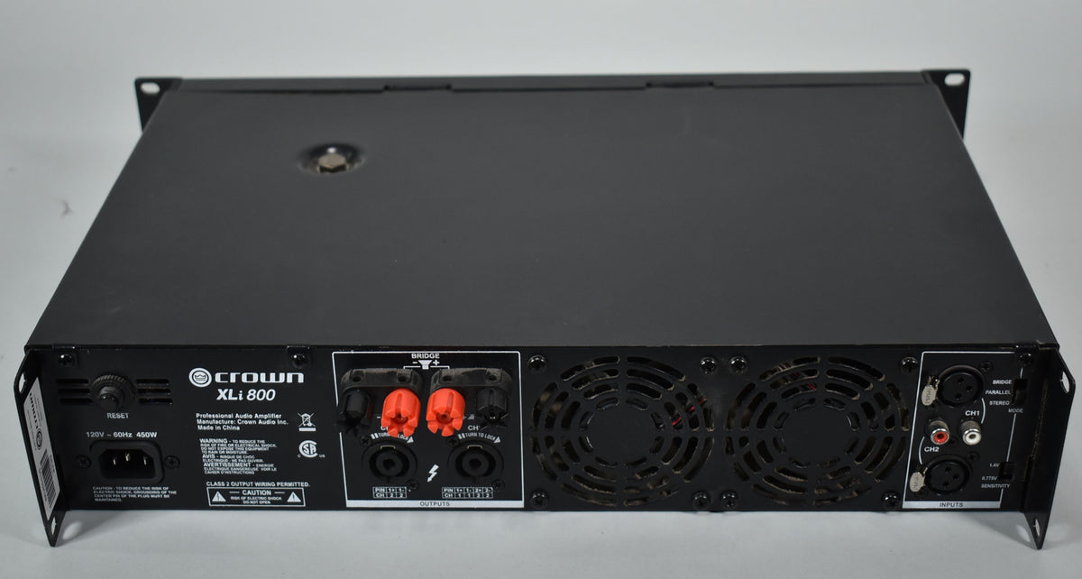 Rear view of a black power amplifier, showing two large cooling fans, red and black speaker terminals, and various input jacks.