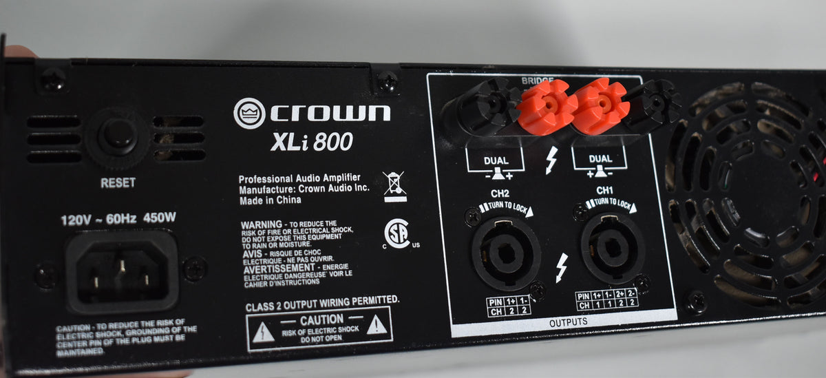 The black metal amplifier's rear panel is shown, featuring a power input, reset button, two red and black binding posts, and two XLR outputs.