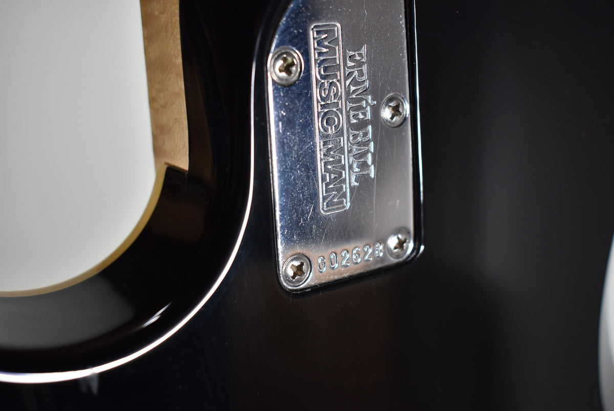 Close-up of a guitar's chrome neck plate, engraved "ERNIE BALL MUSIC MAN" and serial number "G02628", on a black body.