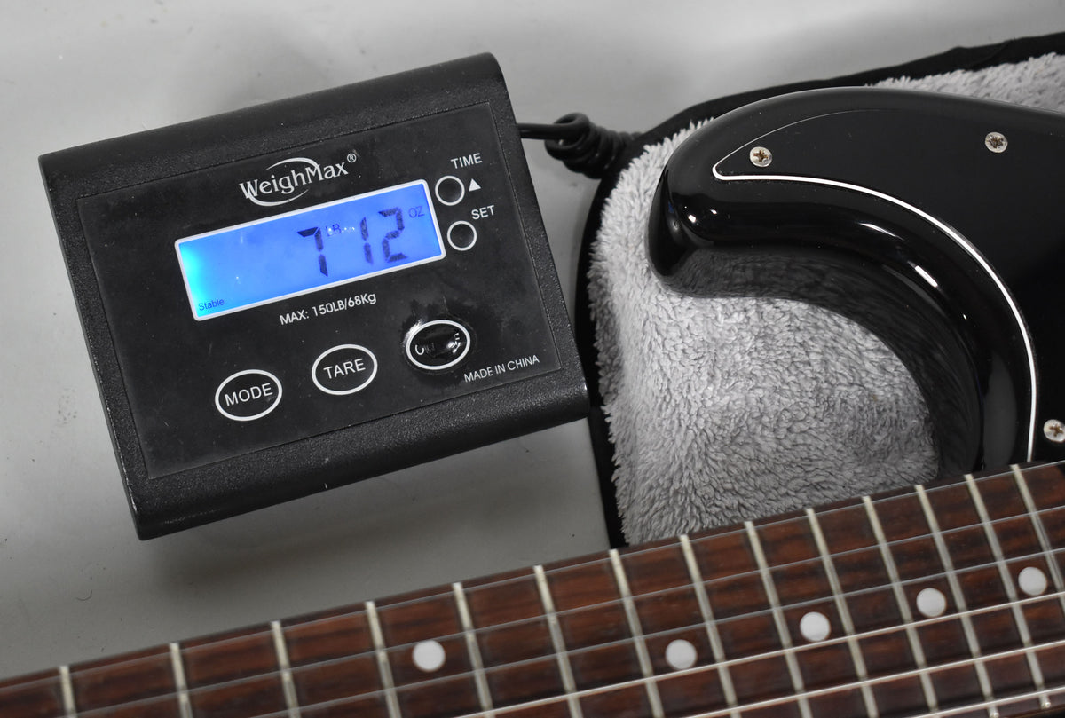 Close-up of a black digital scale displaying "7.12 oz", with the guitar's dark fretboard and part of its black body visible.