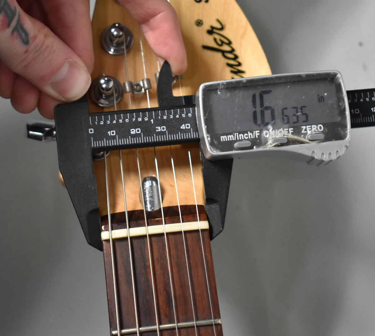 A hand holds calipers measuring the guitar's nut width at 1.635 inches, showing the headstock, tuners, and rosewood fretboard.