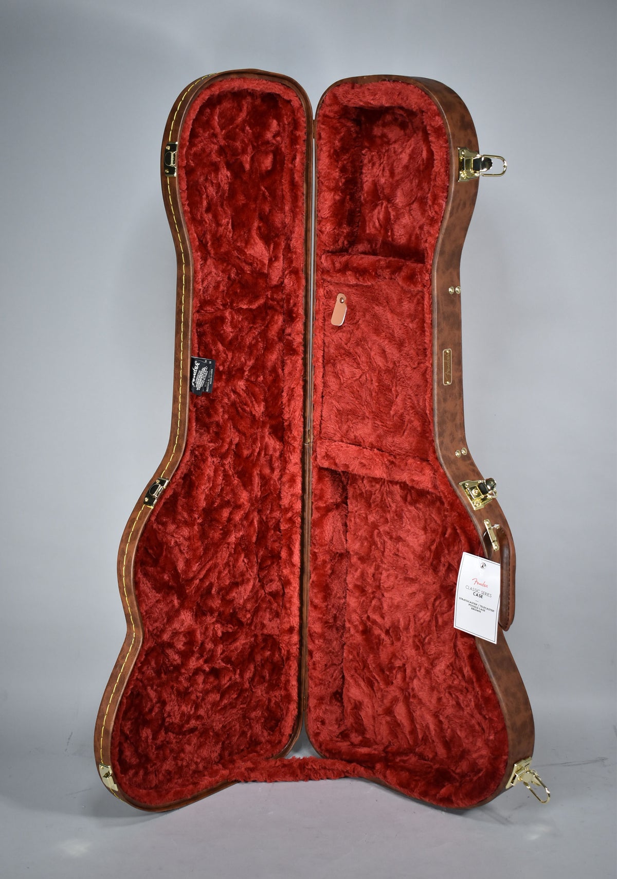 An open brown guitar case with gold hardware and red plush interior, viewed from above, showing an accessory compartment.
