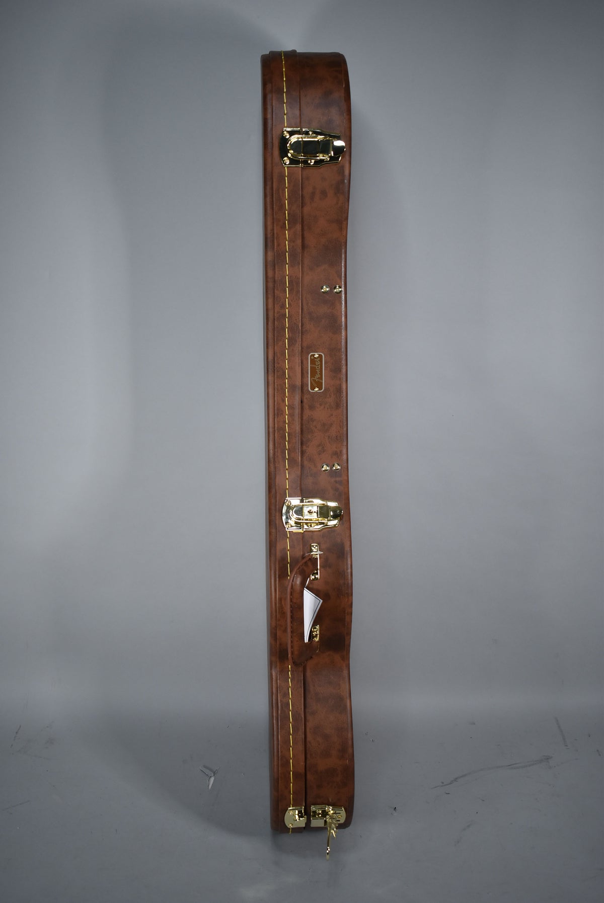 The brown leatherette guitar case is shown from the side, revealing gold latches, a handle, and yellow stitching along the edge.