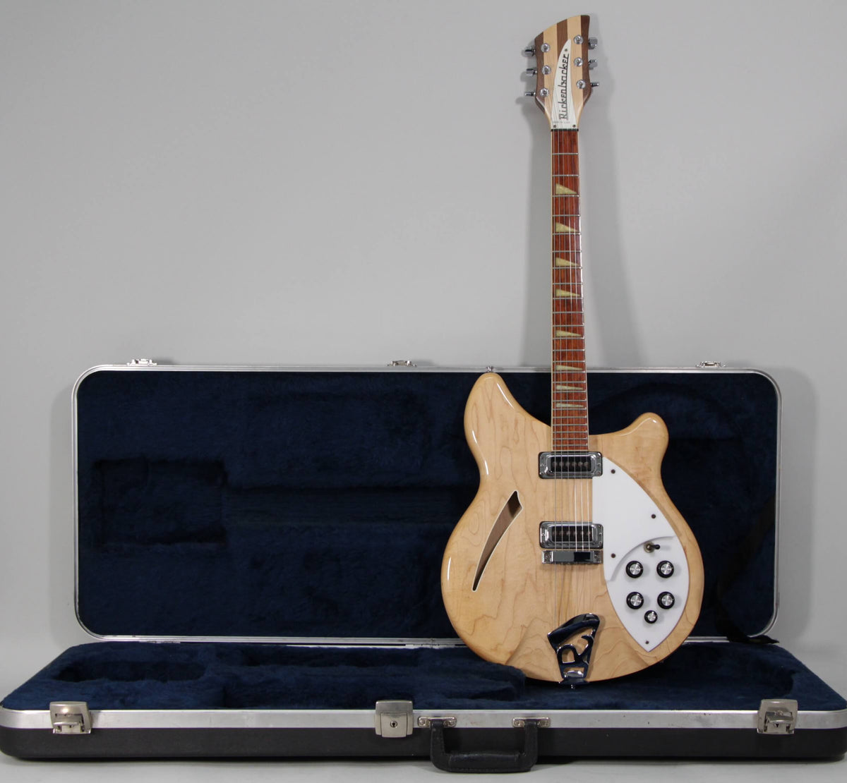 Full-length, natural wood finish electric guitar with white pickguard and five black control knobs, standing in an open blue-lined hard case.