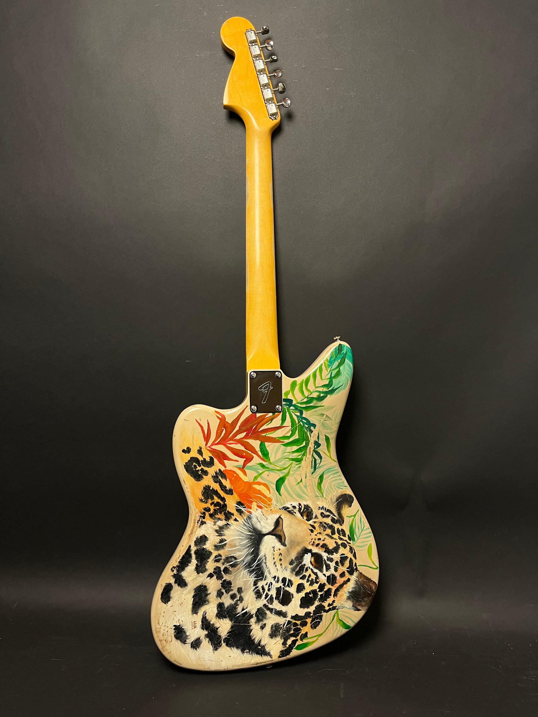 Full rear view of a light-colored guitar with a hand-painted jaguar and tropical leaves, a light wood neck, and chrome tuners.