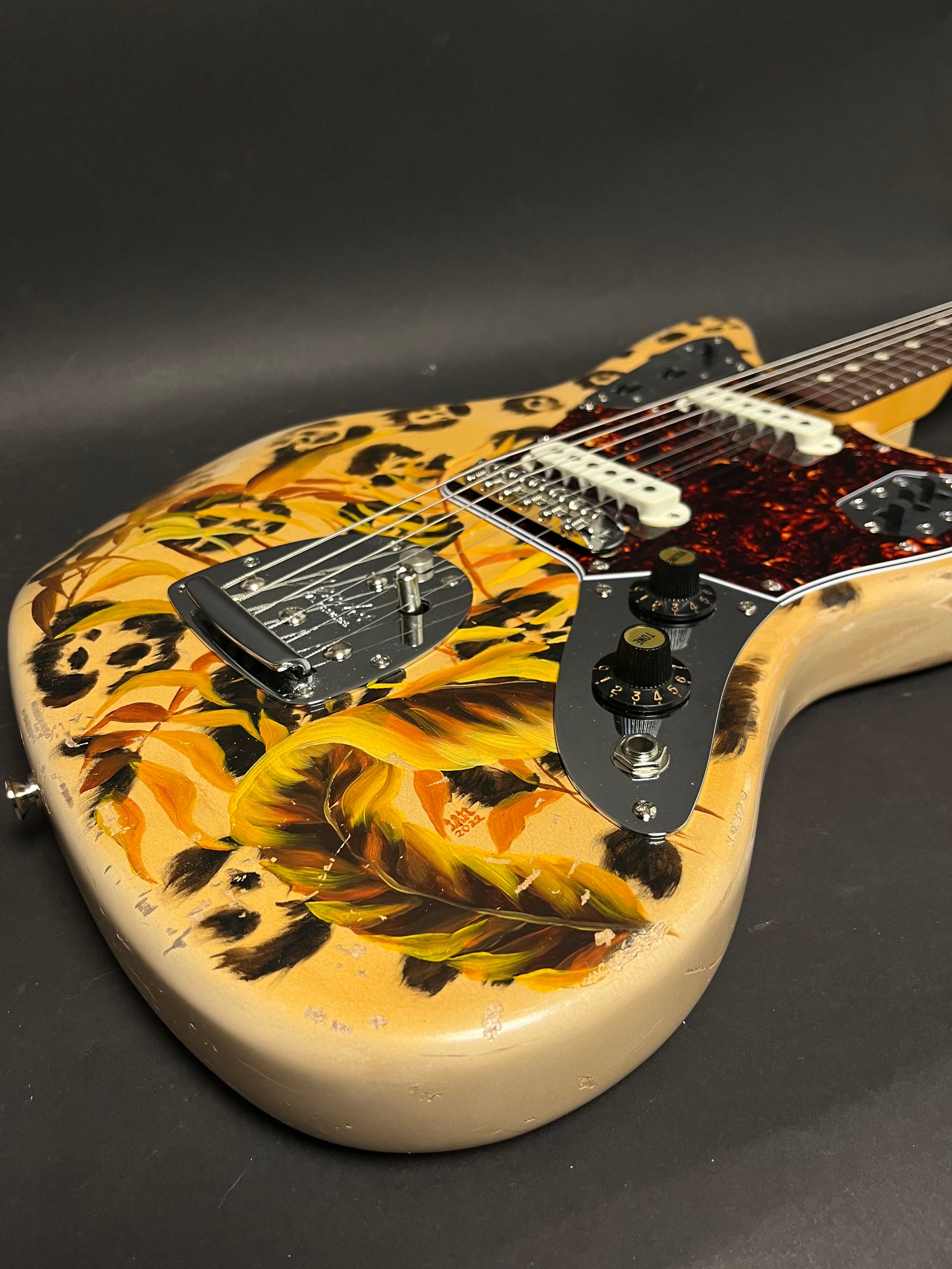 The guitar body, angled from above, shows a leopard print and yellow-orange feather design with visible finish wear and checking.