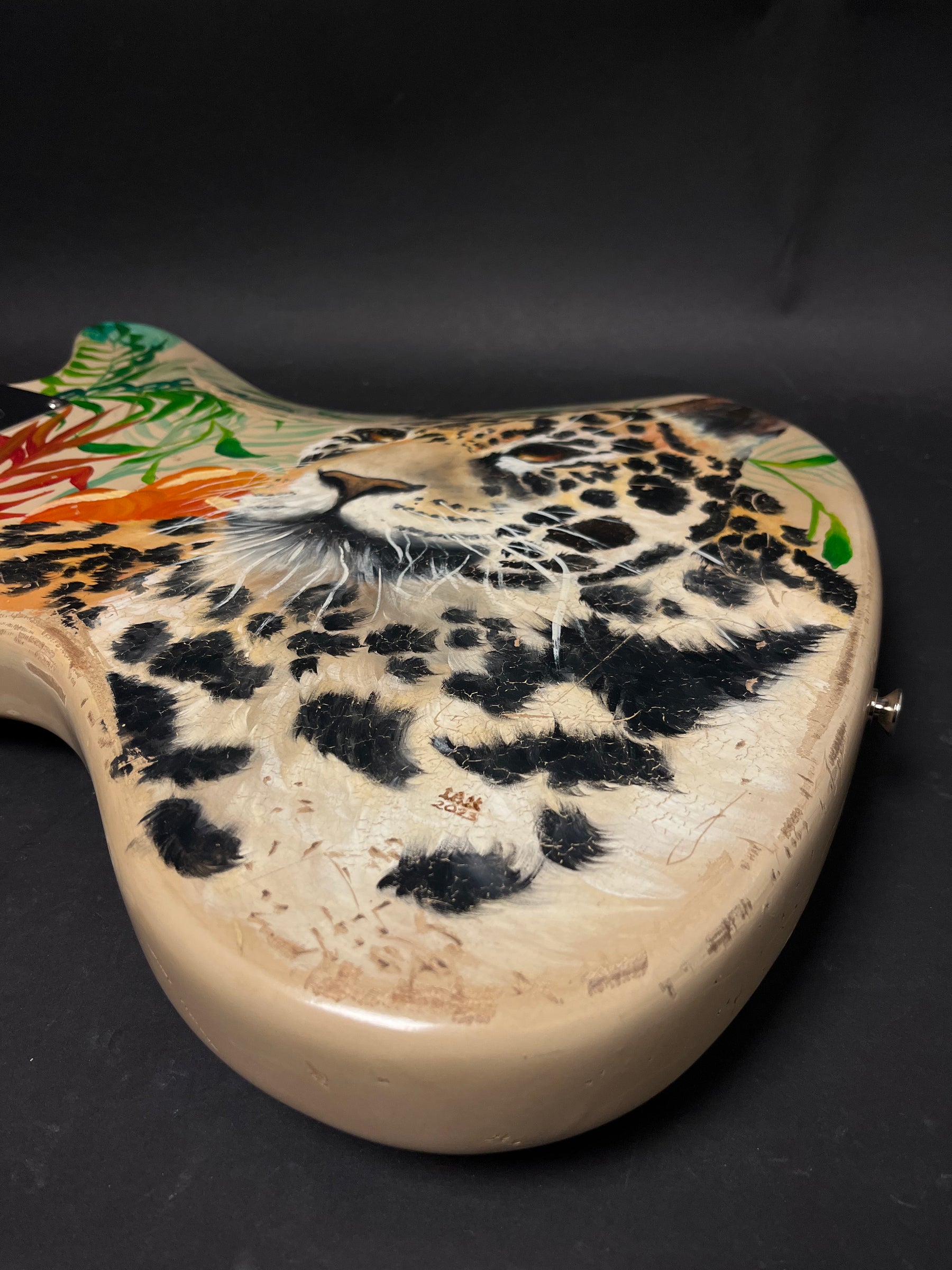 The guitar body, angled right, shows a jaguar's head and spots with tropical leaves, all with a distressed, crackled finish.
