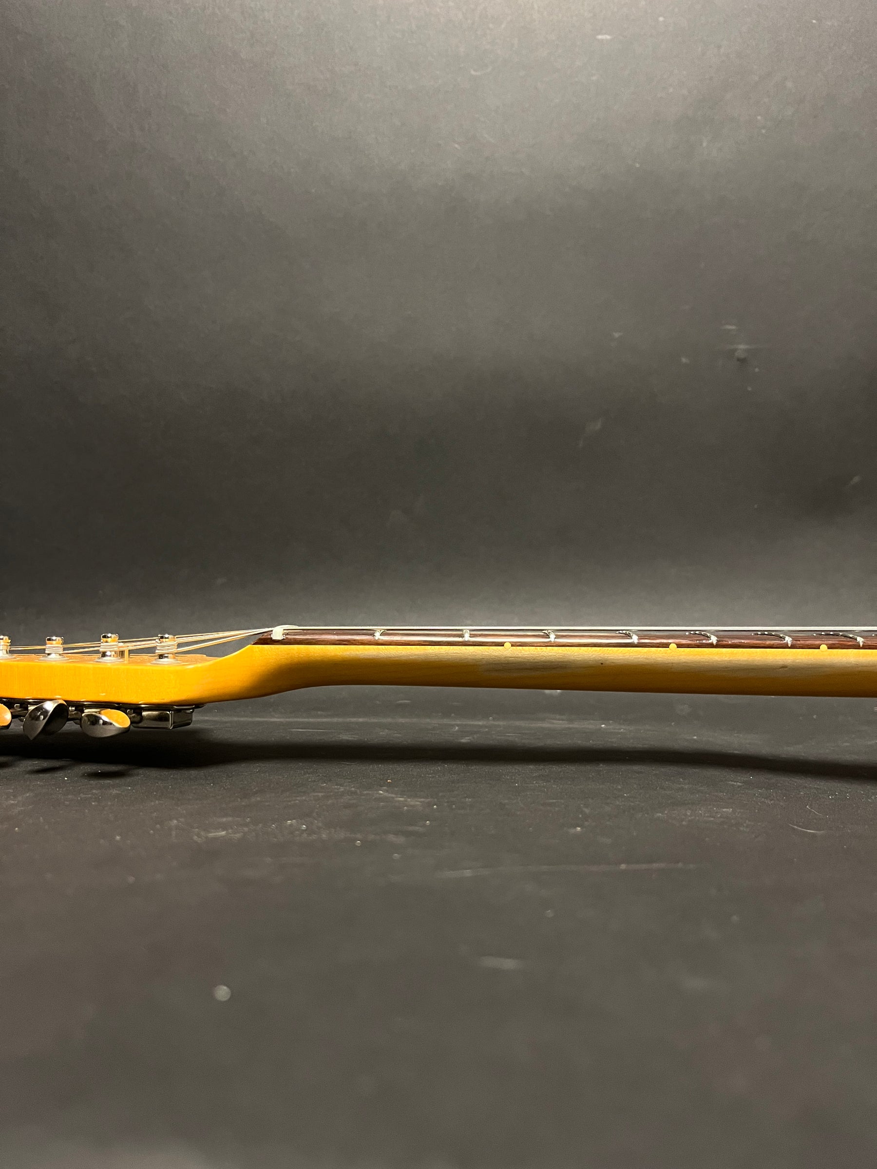 Side view of the guitar neck, showing a light amber finish, dark fretboard with light fret markers, and chrome tuners.