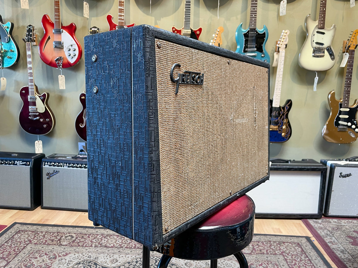 The amp is angled, showing its textured dark blue side and tan grille cloth with a metal logo, on a red stool.