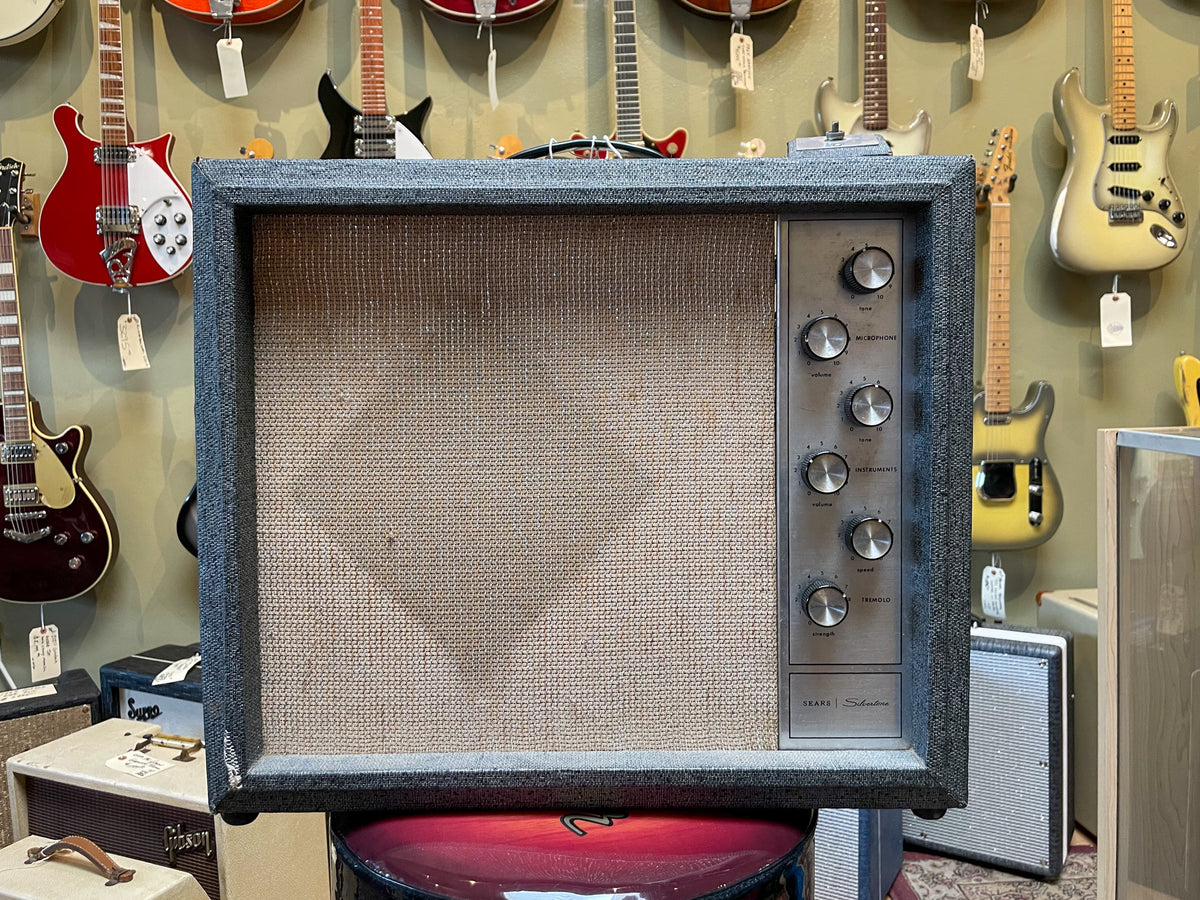Front view of a vintage guitar amp with dark gray tweed covering, a tan grill cloth, and a silver control panel with six knobs.