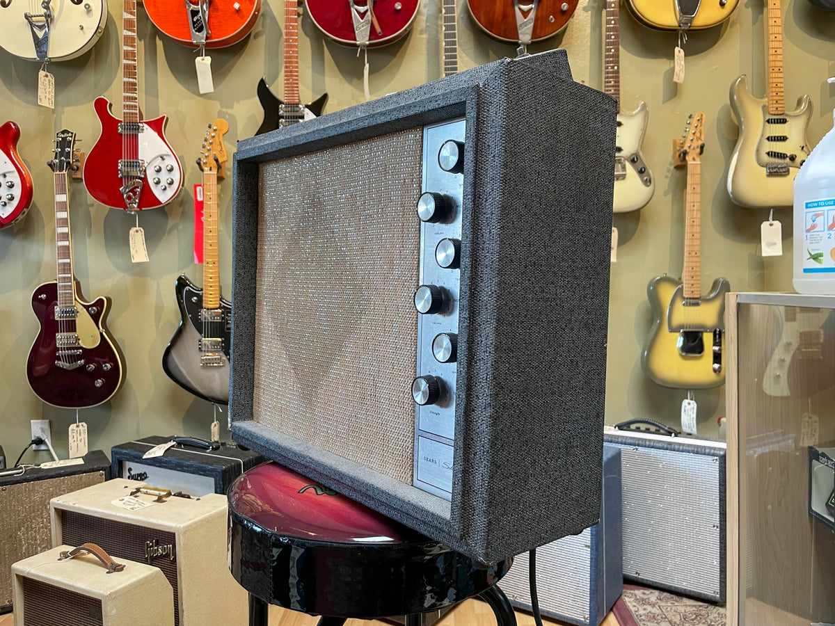 A vintage guitar amp, dark gray with a tan grille, stands angled right, showing its silver control panel with five black knobs.