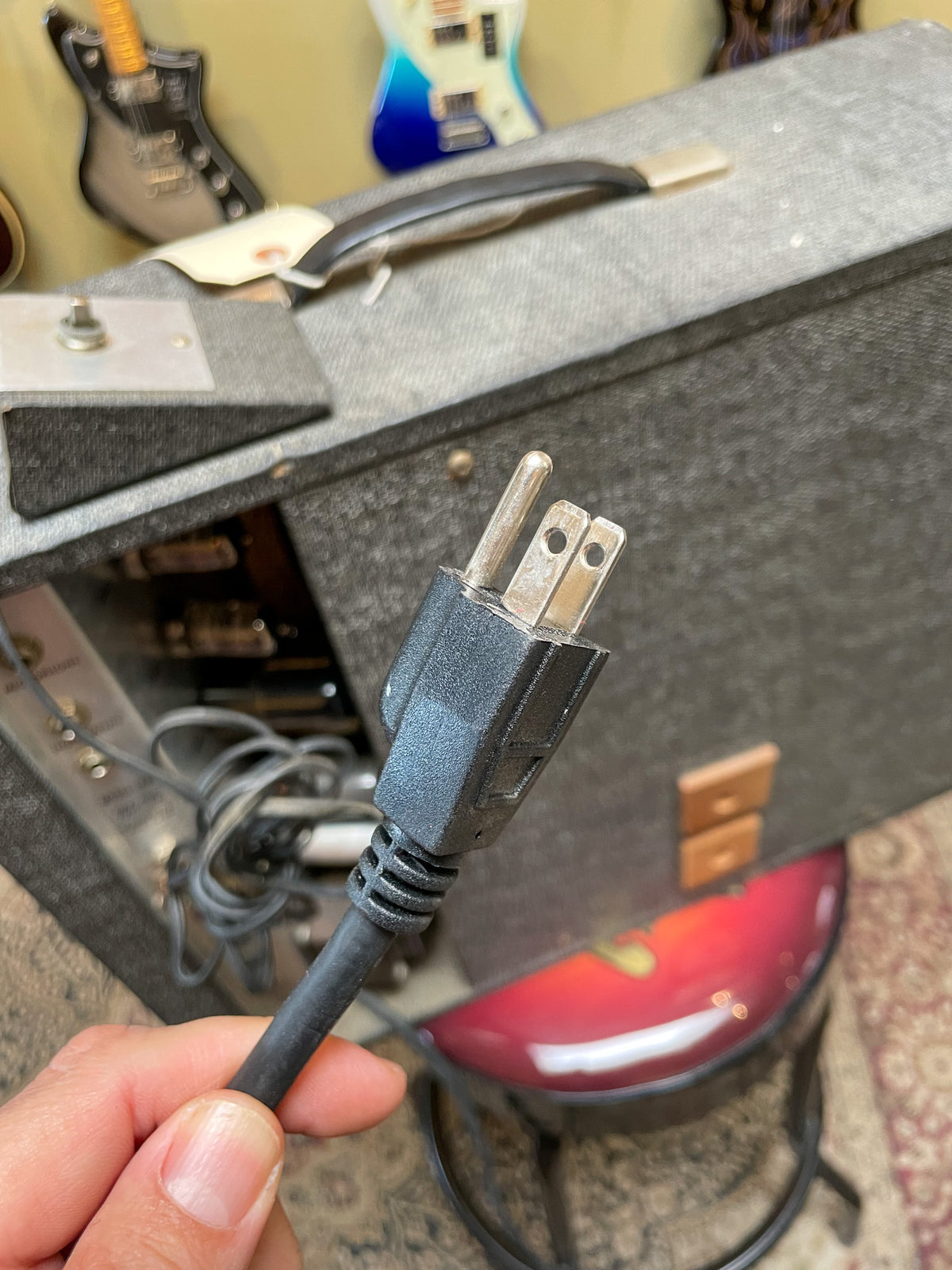 A hand holds the amp's black, three-prong power plug, with the amp's gray tweed cabinet and open back panel visible behind it.