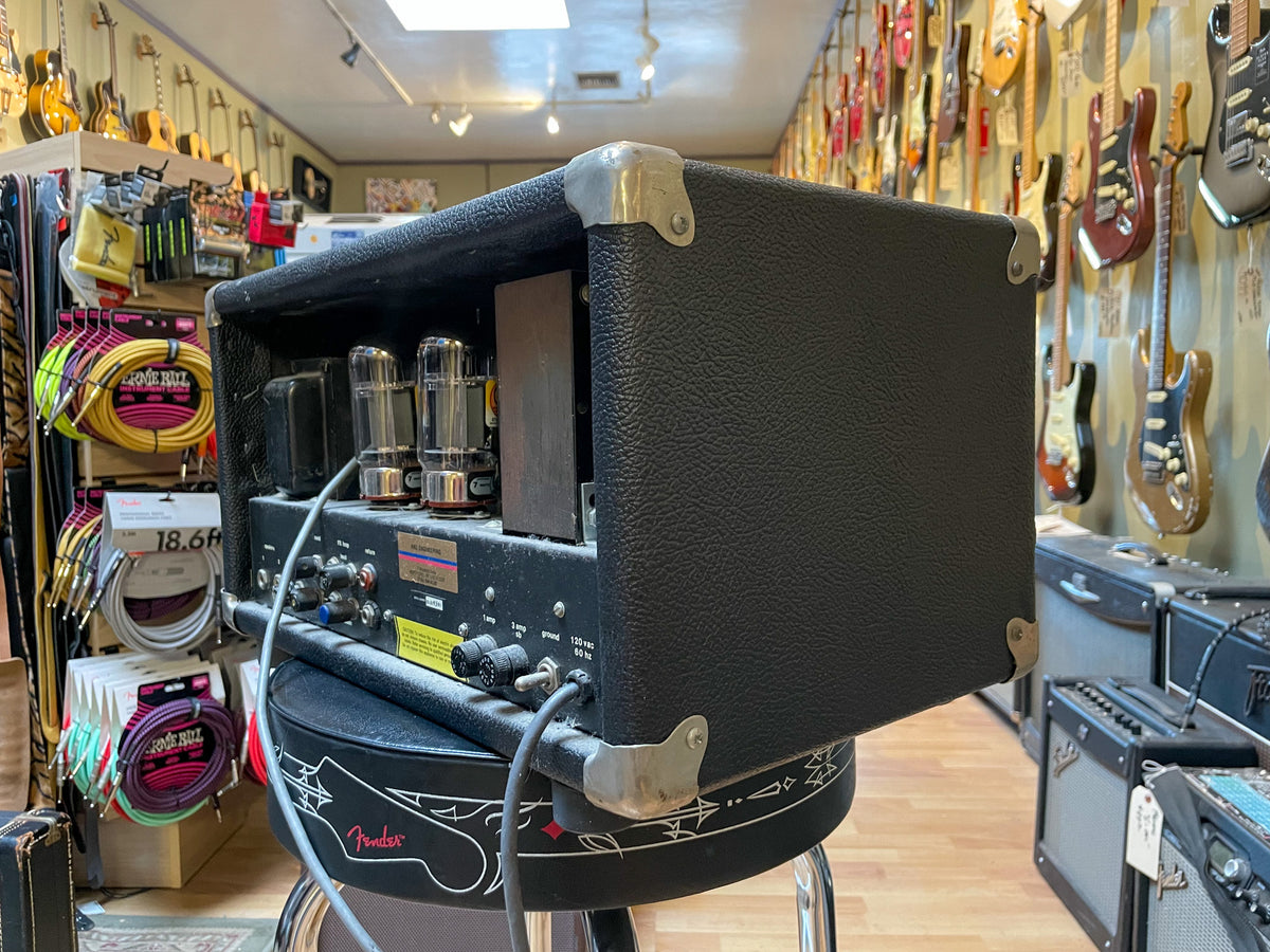 Black textured amp head, viewed from the rear-left, showing exposed vacuum tubes and various input/output jacks and controls.