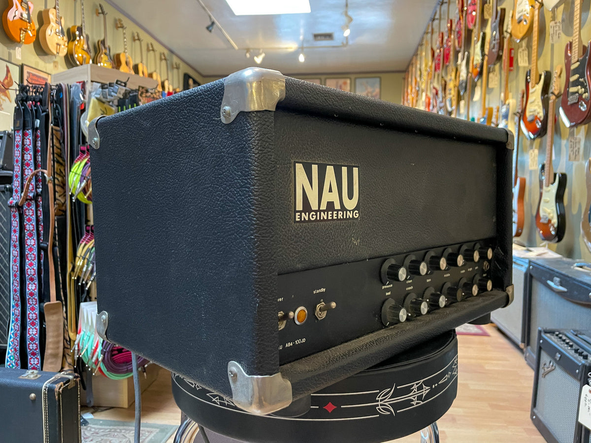 Black textured amplifier head with metal corner protectors, viewed from the left, showing the front panel's many black knobs and two orange switches.