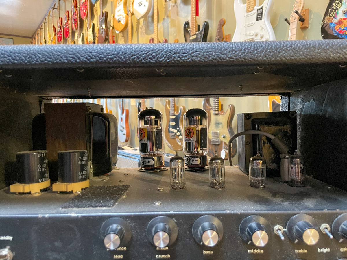 Top-down view of the black amp head's interior, showing four large and four small vacuum tubes, capacitors, and the control knobs below.