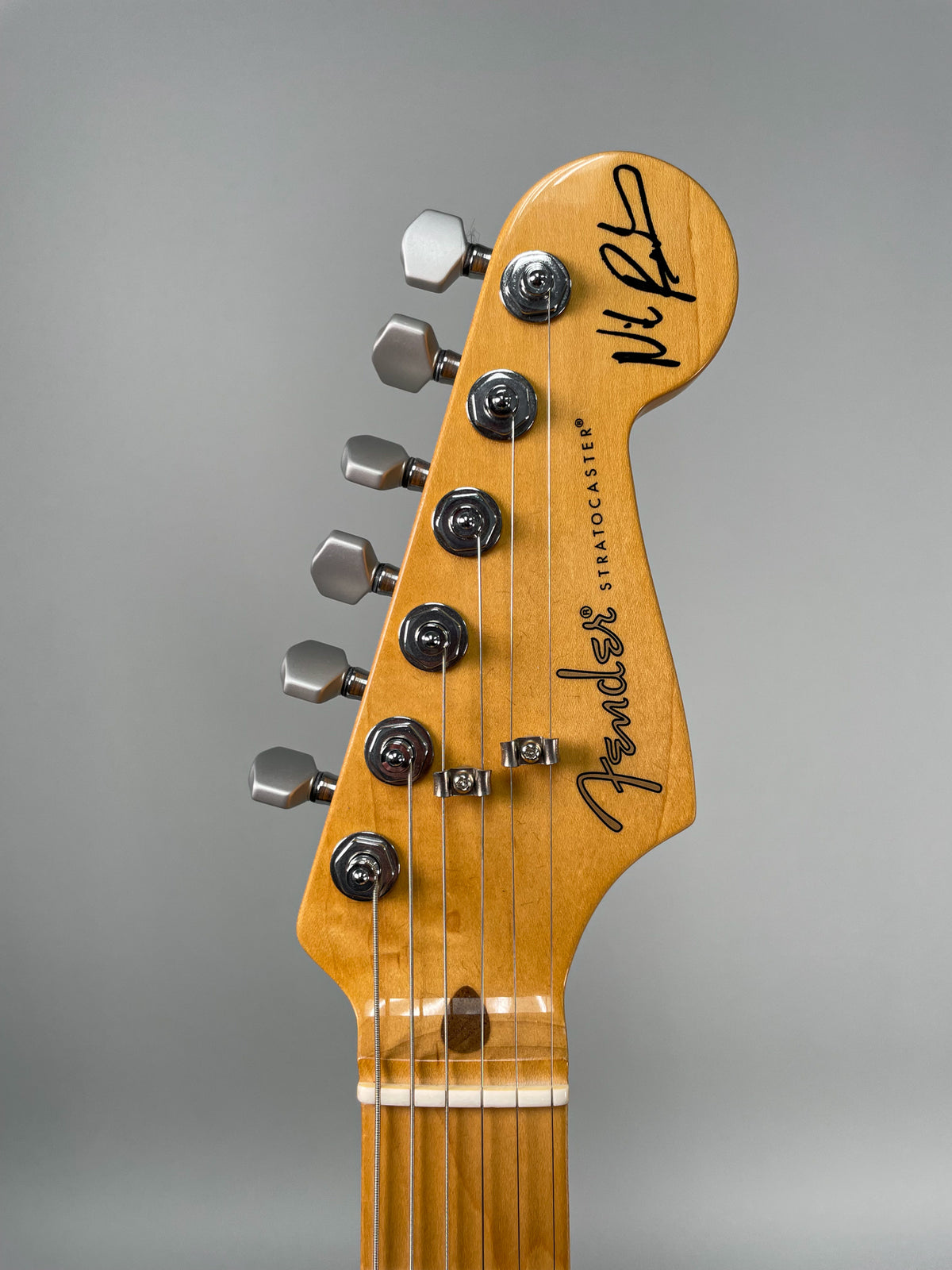 Close-up of the guitar's headstock, angled slightly left, showing six satin-finish tuners and a signature on the light wood.