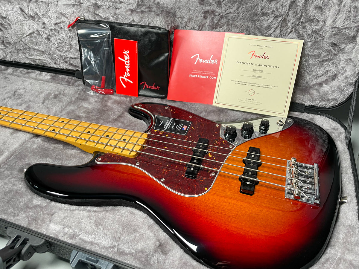 A sunburst Jazz Bass with a tortoiseshell pickguard, maple fretboard, and chrome hardware, shown in its open case with accessories.