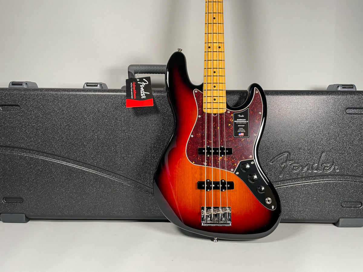 A sunburst bass guitar with a tortoiseshell pickguard and maple fretboard stands upright in front of its black hard case.