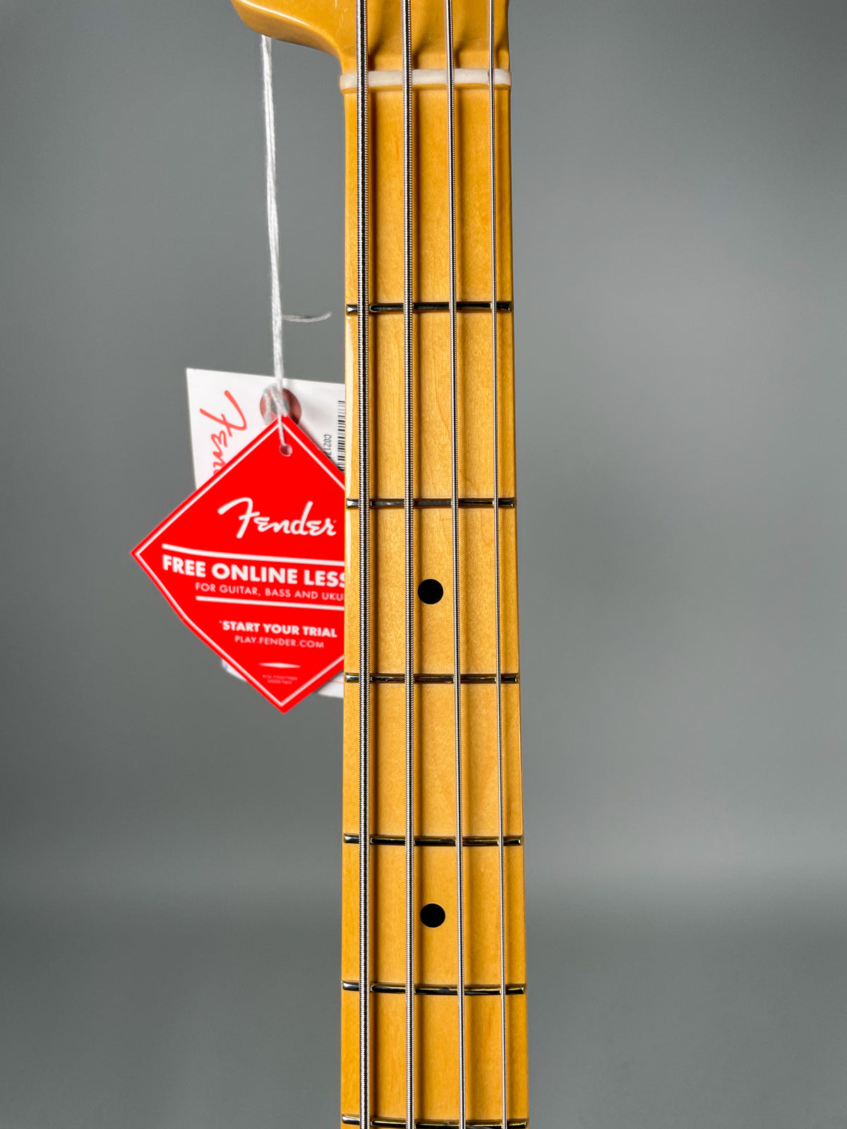 Close-up of the bass guitar's maple fretboard with black dot inlays, showing the strings and a red Fender tag hanging from the headstock.