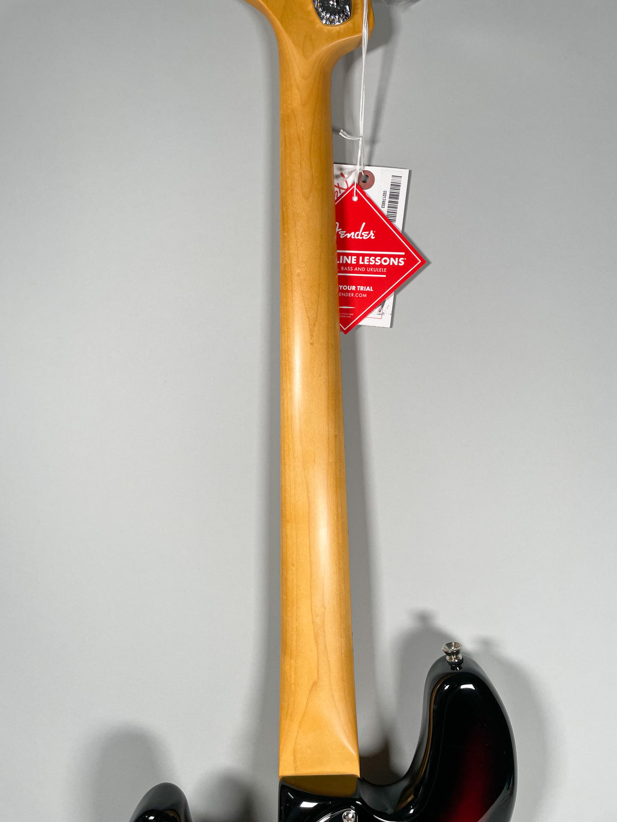 The bass's glossy maple neck and headstock are visible from the back, with a red hangtag near the tuning pegs.