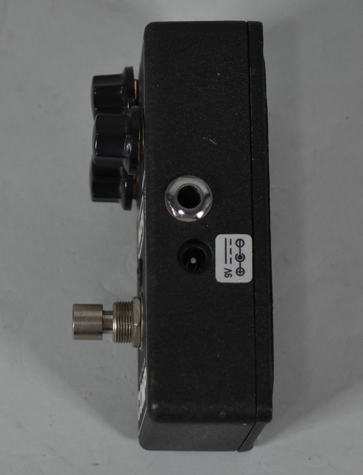 The black textured pedal is viewed from the side, showing three black control knobs, a jack, a power input, and a 9V sticker.