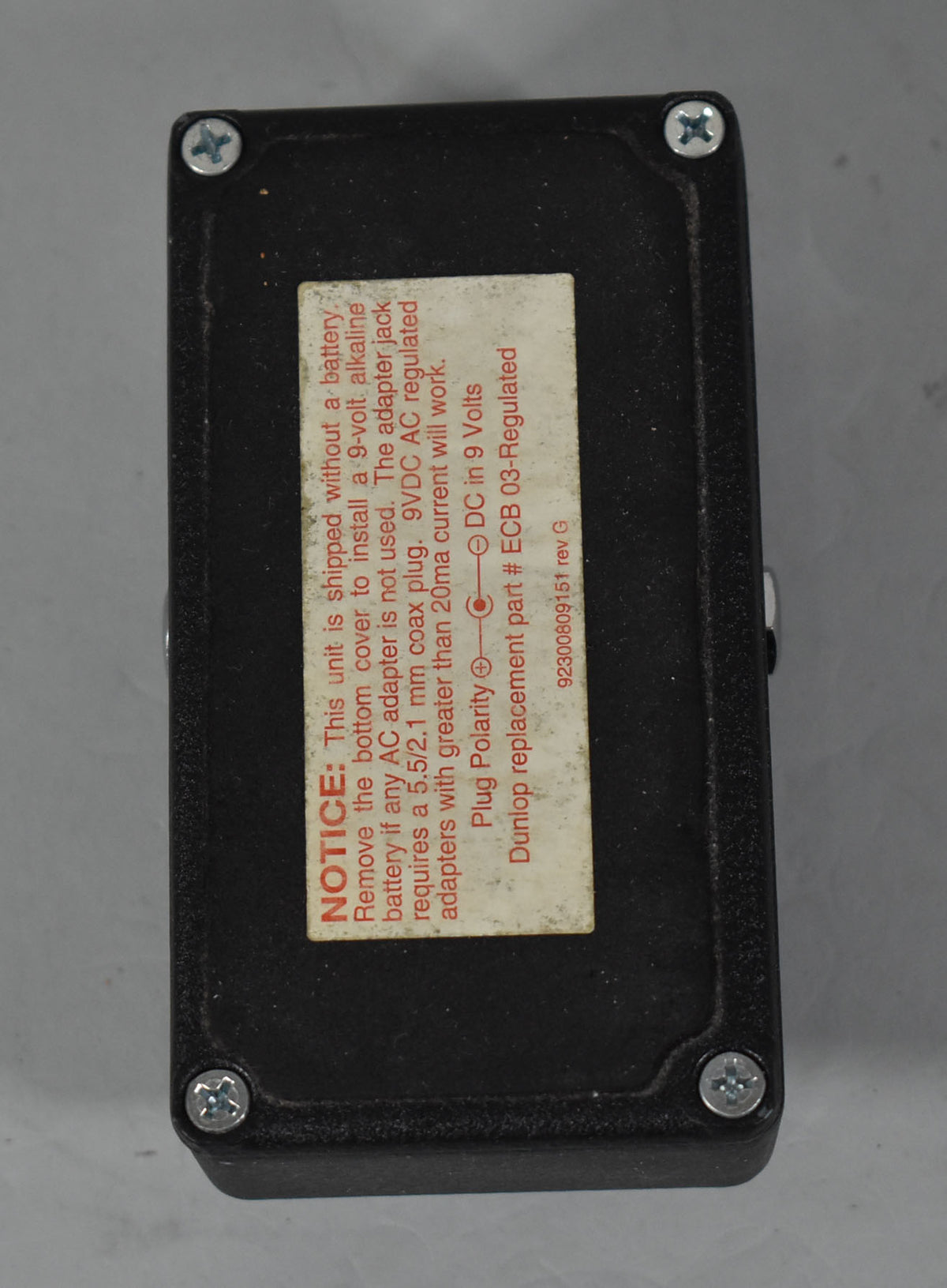 The black metal pedal is viewed from the bottom, showing a large rectangular white warning label with red text and four silver screws.