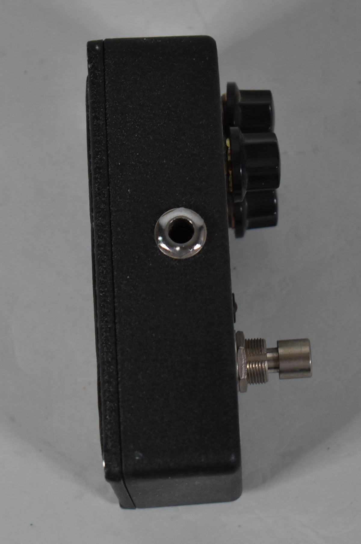 The black textured pedal is shown from the right side, revealing a chrome input jack, three black knobs, and a footswitch.