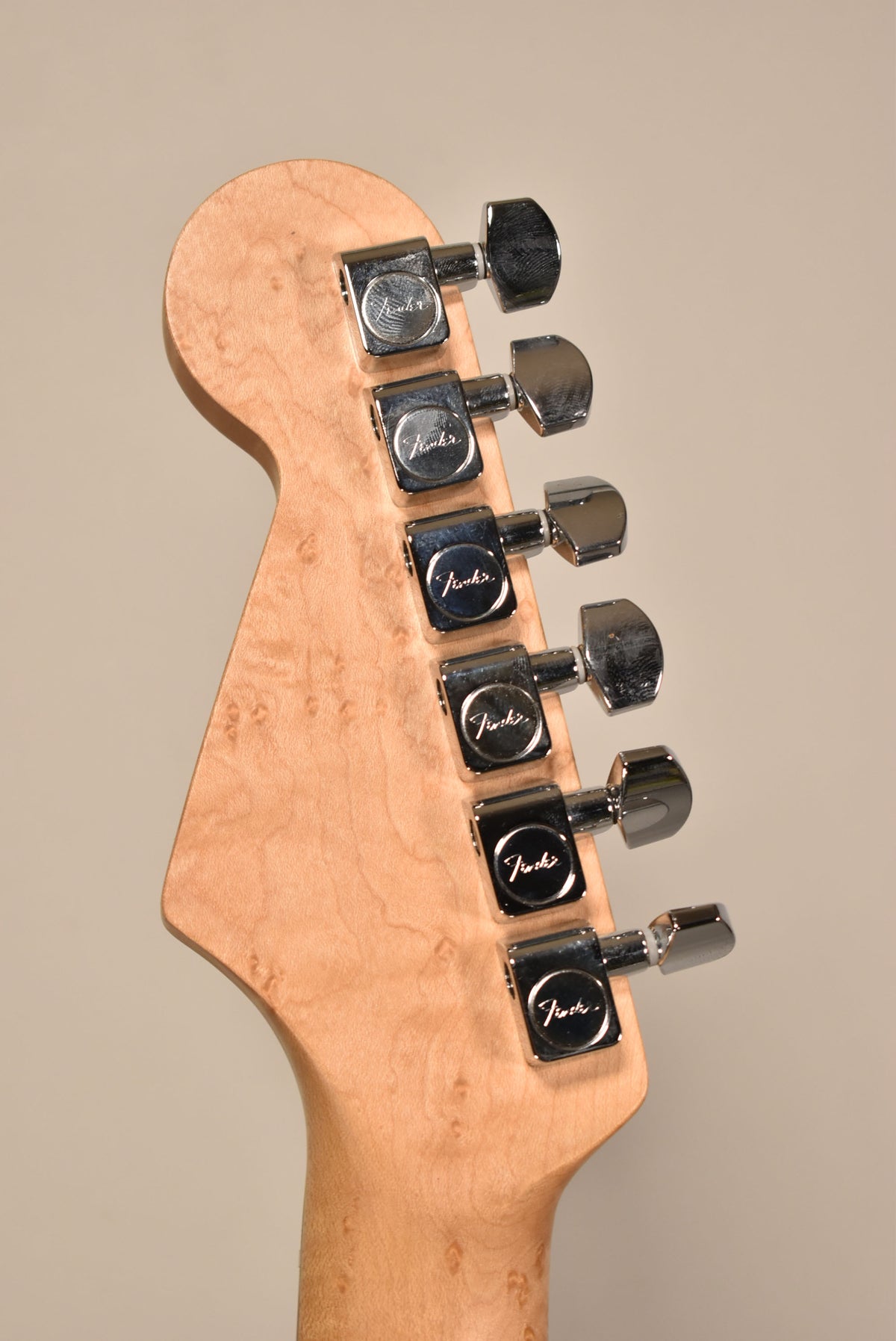 The back of a light-colored, figured maple headstock with six chrome tuning machines, viewed from a slight angle.