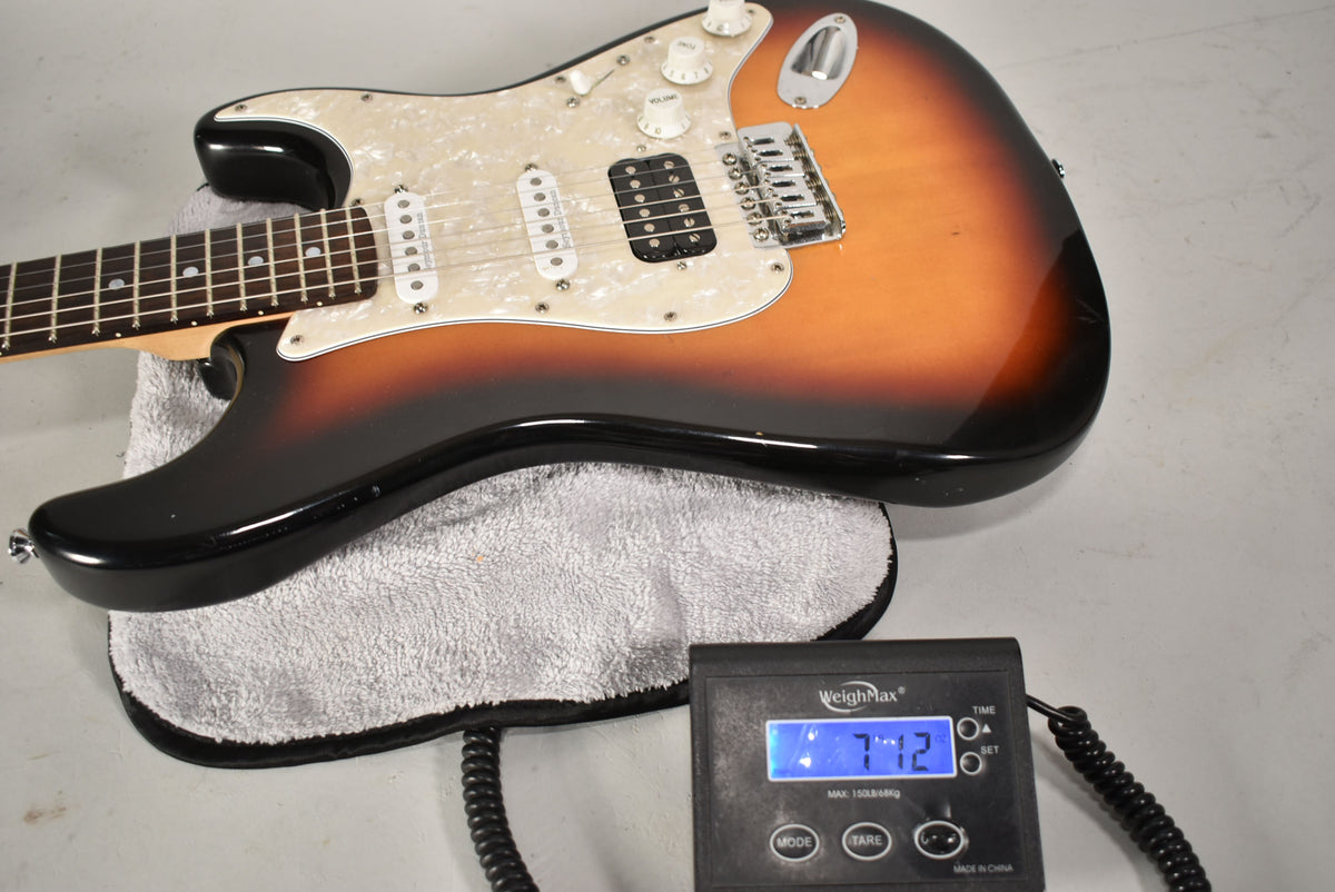 The sunburst Strat-type guitar, with a pearloid pickguard and HSS pickups, rests on a towel, showing a scale displaying "7.72".