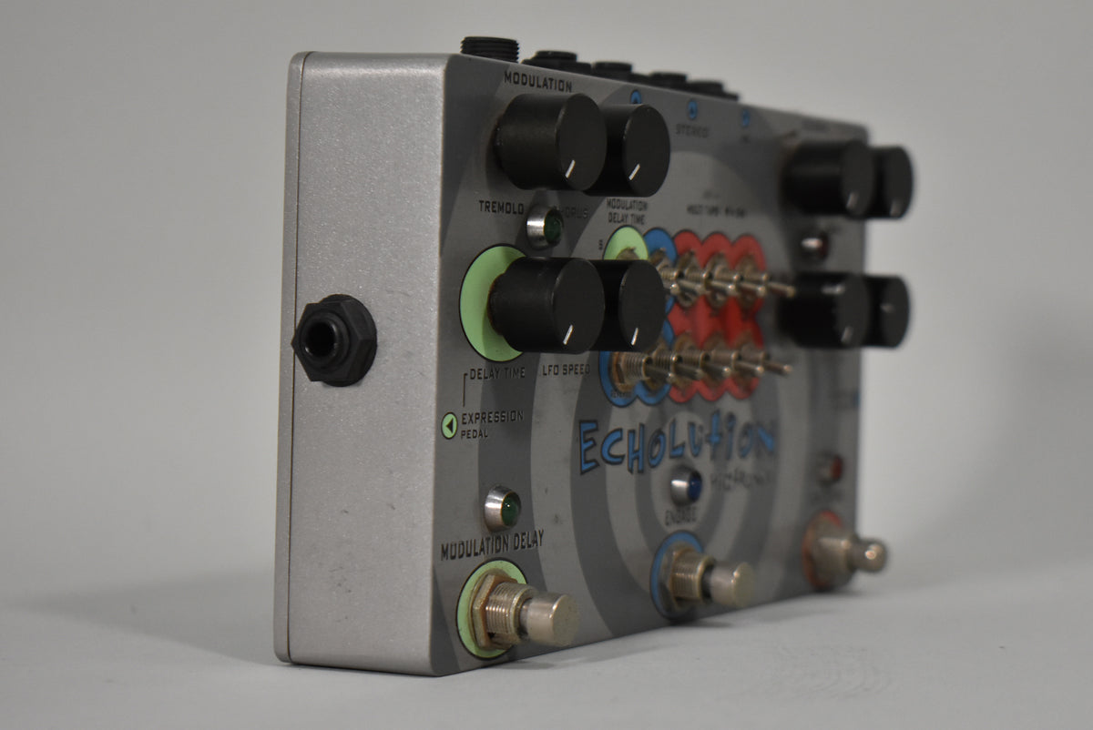 A silver-grey pedal, angled right, shows its left side input jack, four black knobs, and six red and blue toggle switches.
