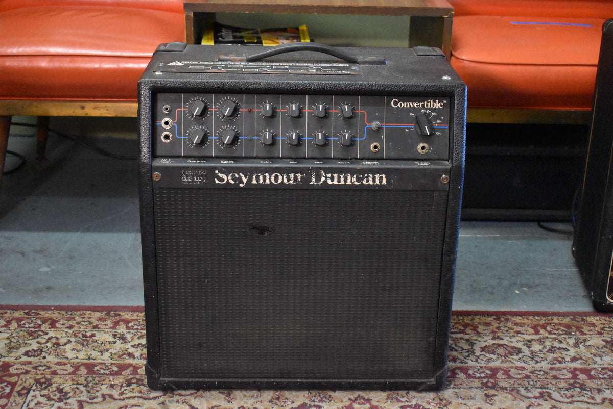 Front view of a black combo amp with a textured grille, 12 knobs, 3 input jacks, and a torn grille cloth.