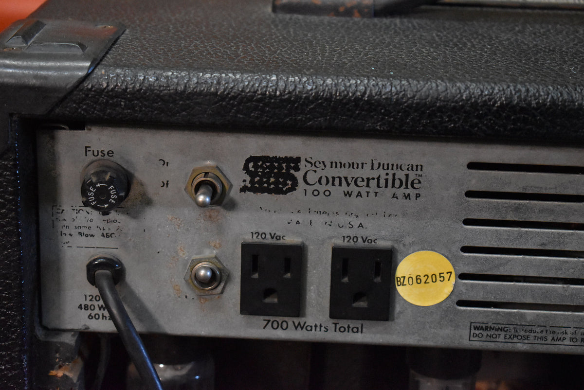 Close-up of the amp's textured black rear panel, showing a fuse, two toggle switches, two power outlets, and a yellow sticker.