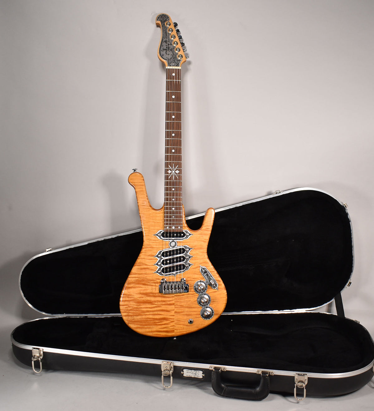 Full view of a natural finish electric guitar with a flame maple top, three humbuckers, and an ornate headstock, resting in an open hard case.