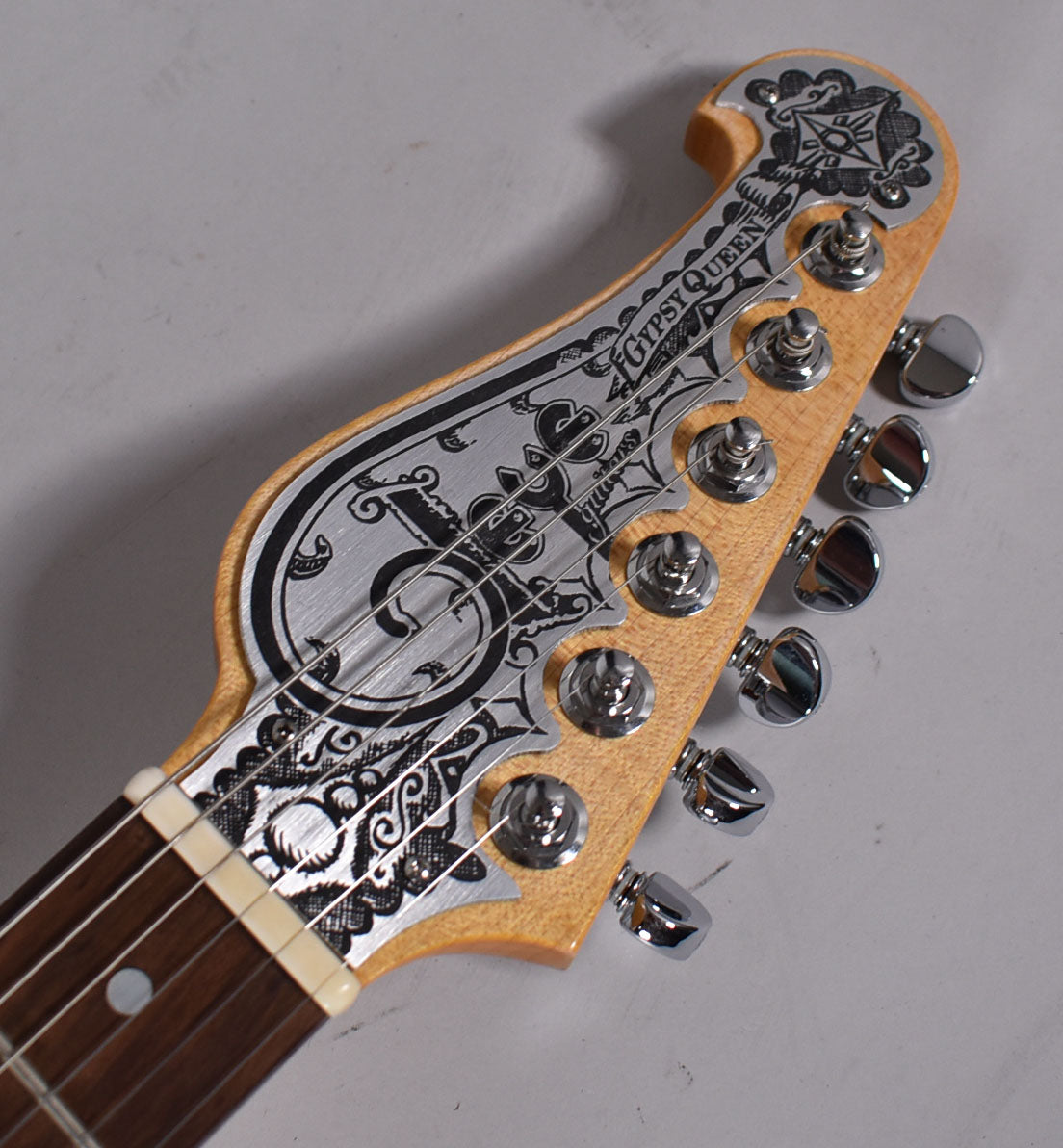 Close-up of the headstock, showing a natural wood finish with an ornate, brushed metal overlay and six chrome tuners.