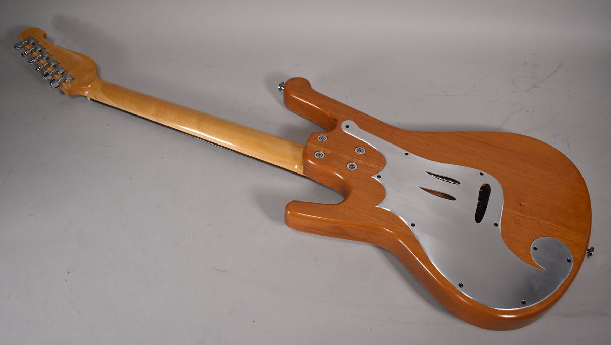 The natural finish electric guitar is angled from the back, showing its light wood body, maple neck, and brushed aluminum pickguard.