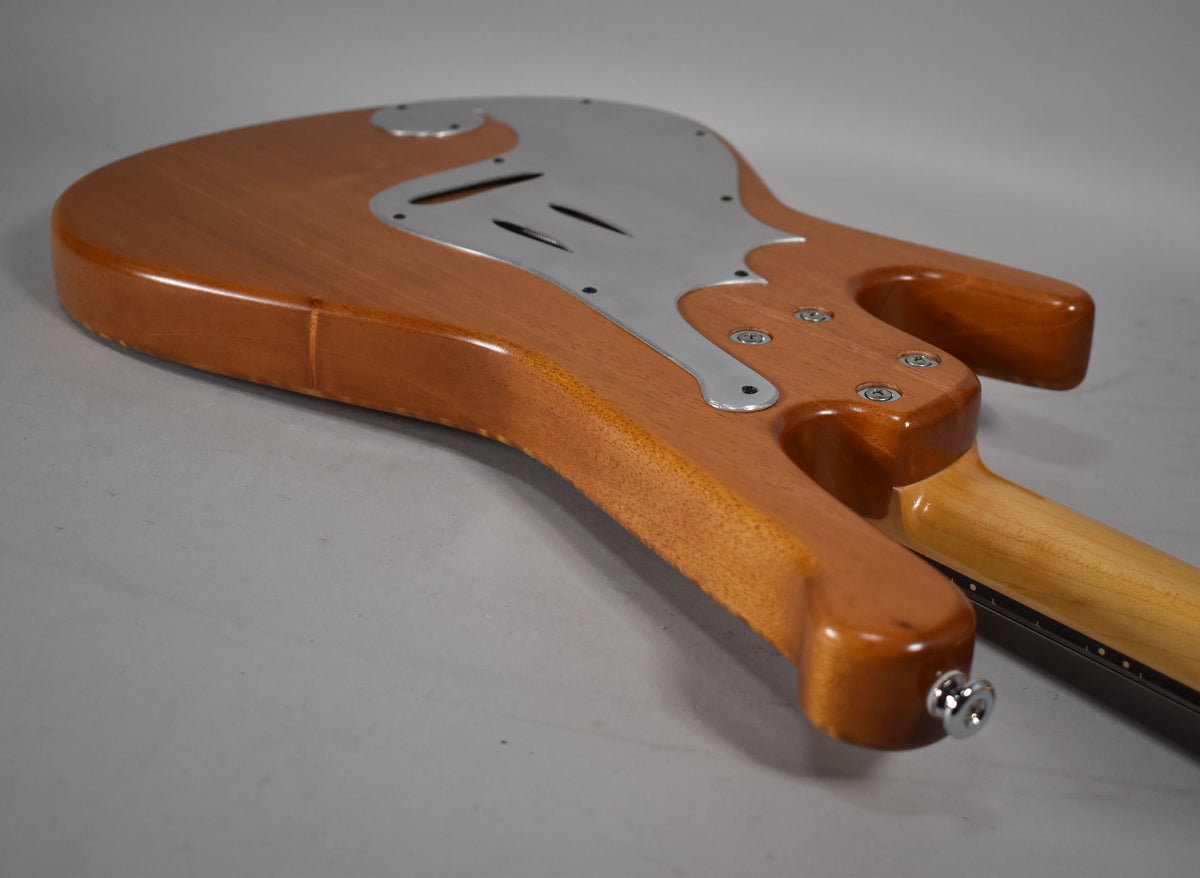 The guitar's natural finish body and neck are seen from the back, with a chrome control plate and output jack.