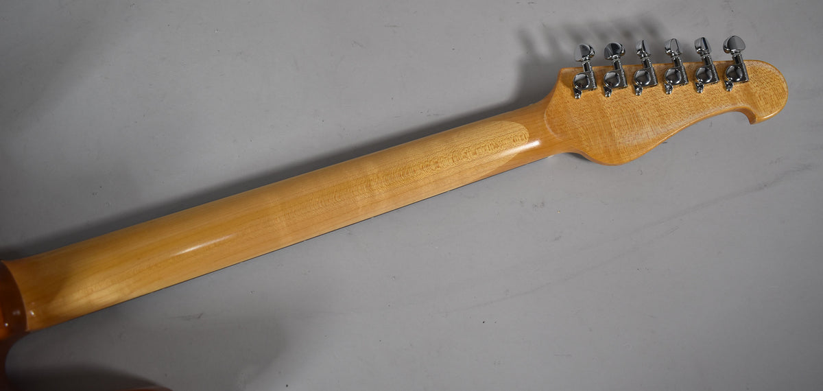 The guitar's light brown natural finish neck and headstock are viewed from behind, with six chrome tuners visible.