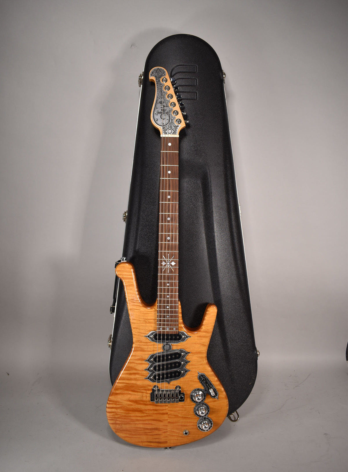 Full view of the natural flame maple guitar with ornate headstock and fretboard inlays, standing upright against its black hard case.