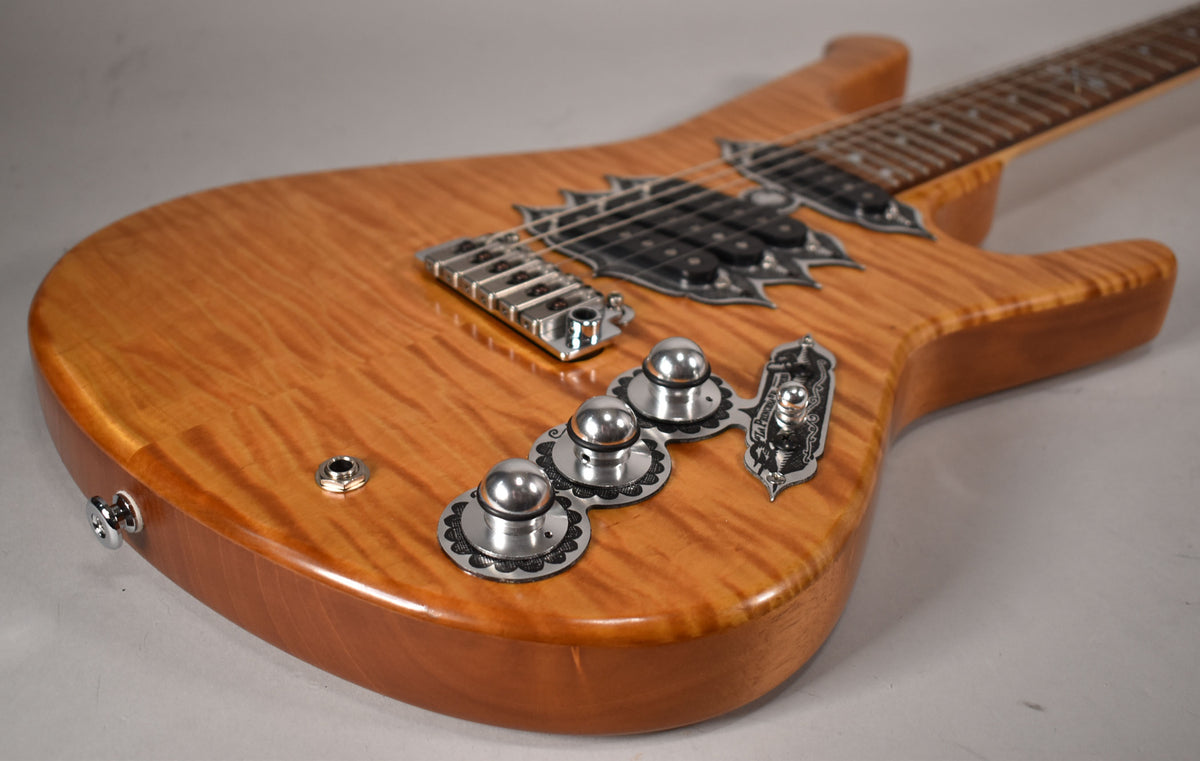The natural finish electric guitar's body, with flame maple top, shows ornate chrome hardware and three dome-top control knobs.