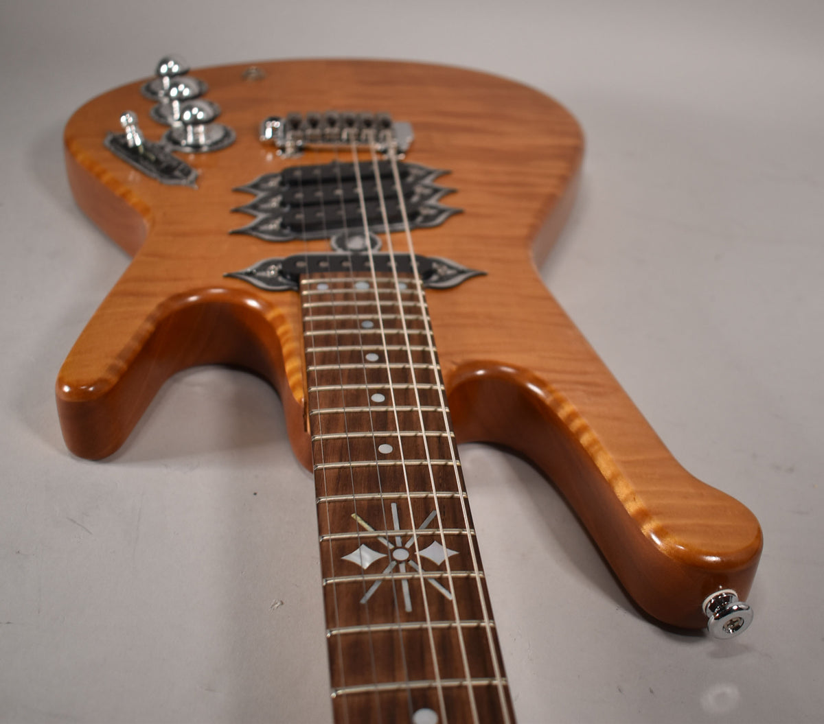 The natural finish electric guitar is angled from the headstock, showing the flame maple body, ornate pickup surrounds, and sunburst fretboard inlay.