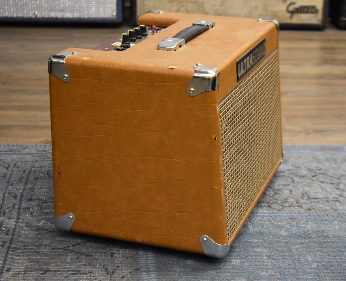 Three-quarter view of a tan combo amp with a gold grille, chrome corner protectors, a black handle, and top-mounted controls.